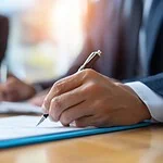 Person signing a document with a pen, with miniature buildings in the background.